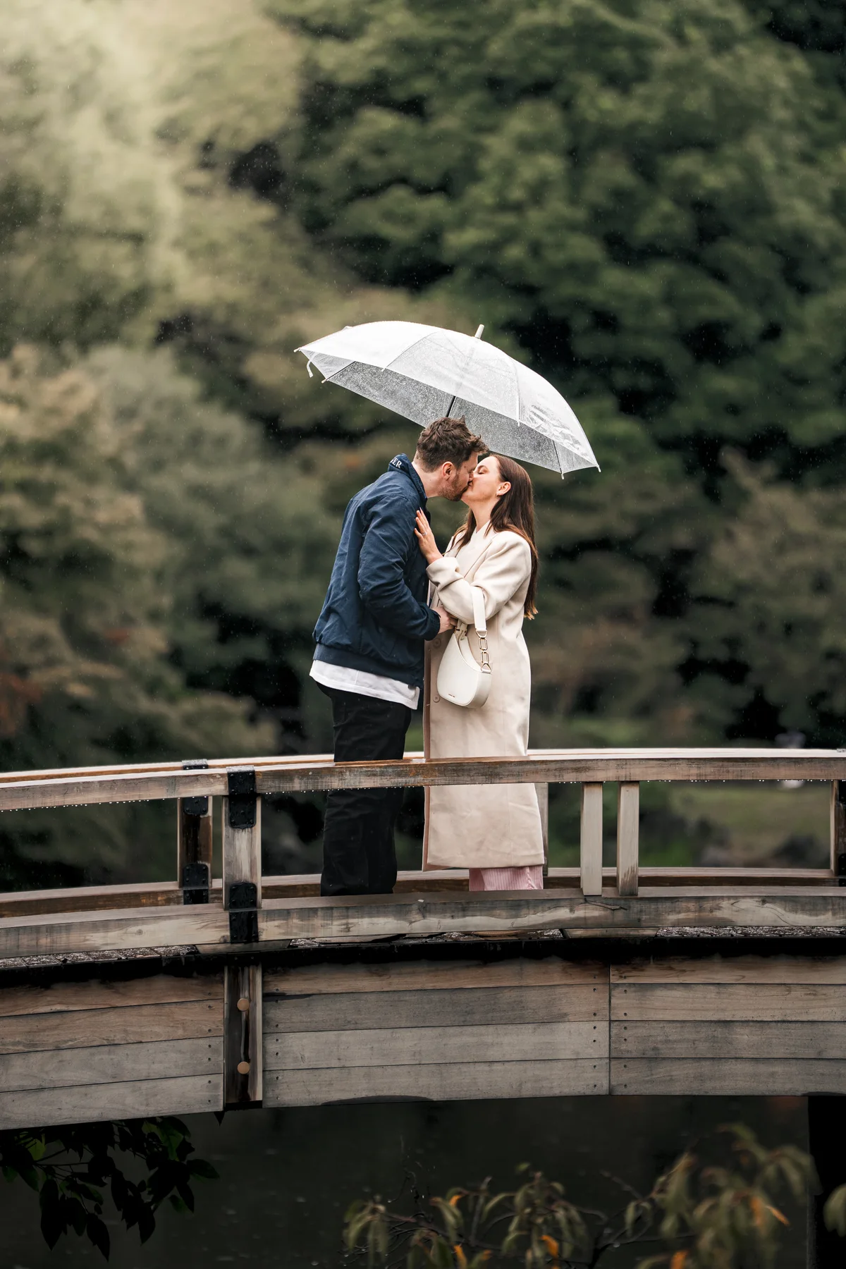 A couple kissing beneath a clear umbrella on a wooden bridge